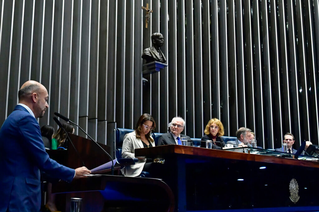 O professor Paulo Soethe discursa no Senado. Ele está no canto esquerdo da foto, falando na tribuna. Também aparece a mesa do senado.