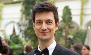 Marcelo Campanér aparece de frente para a foto e com um leve sorriso. Usa um smoking preto com camisa branca e gravata borboleta preta. Ele tem olhos e cabelos castanhos. O fundo da foto está desfocado e se refere ao lançamento de um filme em Cannes.