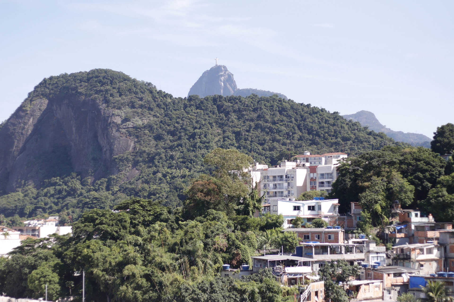 Fotografia do Rio de Janeiro a partir do Leme, em que aparece o Corcovado e o Cristo Redentor ao fundo, e mais a frente aparecem morros com vegetação tropical e a comunidade do Morro da Babilônia.